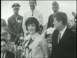 President John F. Kennedy introduces Jacqueline Kennedy to an audience and as she speaks a man interprets in Spanish. News Clip