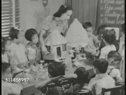 1936: COMMONWEALTH of the PHILIPPINES: PUBLIC SCHOOL: Female teacher w/ Nipa hut model on table surrounded by Filipino kindergarten, or first grade students (poor quality). Children behind craft table. Smiling teacher. Boy holding paper fence & flower Instructional Video