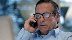 Multitasking businessman scans laptop-computer screen, talks and switches ears on smart-phone in private corporate office (dolly shot) Stock Footage