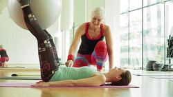 Personal trainer guiding woman squeezing fitness ball between legs in gym Stock Footage