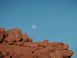 WS T/L View of Clear blue sky ghostly moon sinks down behind earth red rocks as sun lights them up from in front / Spitzkoppe, Windhoek, Namibia Stock Footage