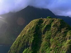 AERIAL aircraft point of view toward summit of hill in ocean with man flying kite / mountains in background / Hawaii Stock Footage