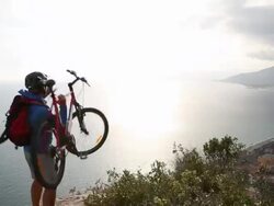 Mountain biker on cliff above sea, raises bike overhead  Stock Footage