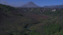 A volcano beyond a green valley spews ash. Stock Footage