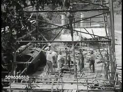 1940: EAST INDIES OIL: WS Refinery tanks on hill. Large crowd of Indonesian people outside building. VS People standing in line. Men working in well area. VS Refinery tanks, buildings w/ smoke stacks, round & oblong tanks. Three round tanks. Gas, oil. Instructional Video