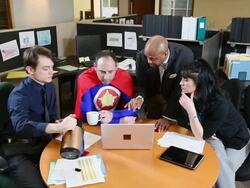 MS Three business people and one man dressing as superhero in blue outfit and red cape share serious discussion and looking at laptop screen / Seattle, Washington, United States  Stock Footage