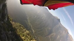 POV from wingsuit flyers foot as he flies over houses, cliffs and forest Stock Footage