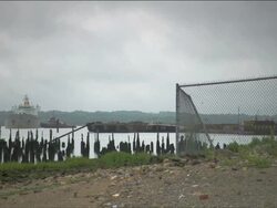Industrial east-coast waterfront with old wooden pilings rising from the water, a chain link fence, and a tanker ship in the distance Stock Footage