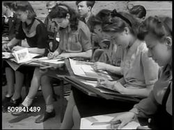 1945: ENGLAND ARCHITECT CLASS: EXT Young British adults w/ pads around city model, instructor at blackboard stand. VS Young students taking notes. MS Instructor talking, blackboard stand, ruined building BG. MS Female student pointing to city model. Instructional Video