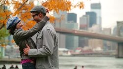 Girl kisses boyfriend's forehead as they embrace in autumn park with Brooklyn Bridge and New York skyline in background (dolly-shot) Stock Footage