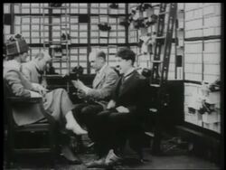 B/W 1916 shoe salesman Charlie Chaplin tickles woman's feet, pushes man, takes shoes from shelf Stock Footage