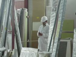 PAN TS man dressed in lab coat and hard hat walking through furniture storage area, checking items and making notes on handheld electronic device Stock Footage