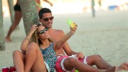 Young Brazilian couple pose for smartphone selfies under palm tree on Copacabana Beach Stock Footage