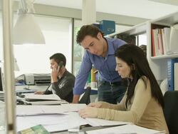 MS male and female office workers sitting at desk, third one (man) standing, discussing work with female office worker in foreground; man in background makes phone call Stock Footage