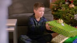 Excited young boy sits down in front of Christmas tree, shakes present and listens intently (dolly-shot) Stock Footage