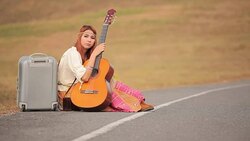 Young hippie woman with guitar and backpack sitting on a countryside road Stock Footage