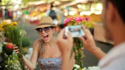 Cute girl hides face behind bouquet of flowers and peeks out for smartphone photo in sunny marketplace Stock Footage