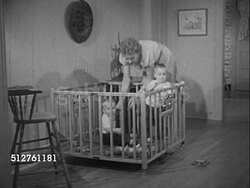 1953: ONE YEAR OLD: Mother carrying twin baby boy into room w/ brother already in wooden playpen, placing baby in pen. CU Baby boy smiling, lifting  & lowering Fisher Price 'Quacky Family' toy, mother duck w/ 3 baby ducks. Instructional Video