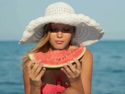 Attractive blond woman in hat eating watermelon on beach Stock Footage