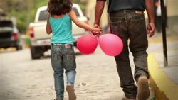 Young girl with balloons holds hands with father while walking up cobblestone street, turns to look at camera Stock Footage