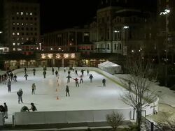 Busy outdoor ice skating rink in city surrounded by tall buildings at night - pan Stock Footage