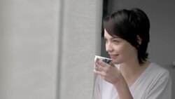 Young woman with morning coffee walks to window and looks out, turns to look back at bed Stock Footage
