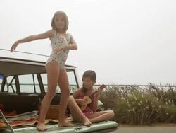 Two girls on car boot, one dancing the other playing guitar Stock Footage