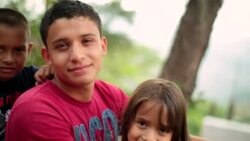 Hispanic teenage brother and younger siblings smile at camera, woman crosses in background Stock Footage