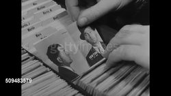 1937: U.S. SECRET SERVICE: MS Two agents at filing cabinet, looking through files. CU Agent flipping through, pulling out mug shot of criminal. MS Man, counterfeiter, in dark cellar at work, making fake prints of dollar bill. Instructional Video