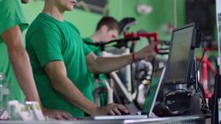 Bike shop employee points at computer screen and explains transaction to coworker at sales counter Stock Footage