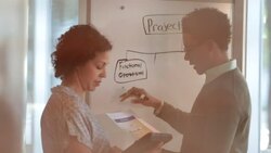 Hip young businessman writes on white-board while female colleague takes notes on tablet, two office workers cross in background (dolly shot) Stock Footage