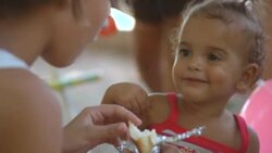 Young Brazilian girl eats sandwich and feeds baby sister in park Stock Footage