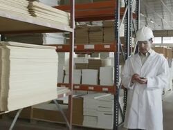MS TS man in lab coat and hard hat walking along multi-story warehouse in furniture production, taking notes on handheld electronic device Stock Footage