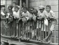 A large group of ethnically diverse children in tattered clothing lean on a porch railing and smile as women rinse laundry in outdoor sinks. Stock Footage