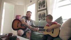 Father teaching son playing guitar on living room sofa Stock Footage