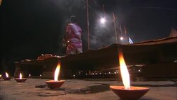 A Hindu monk raises a platter of candles above his head as diyas flicker on the ghat during Diwali. Stock Footage