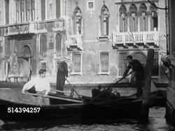 1953: VENICE, ITALY: WS Narrow canal w/ gondolas passing others under arched foot bridge, navigating at turn, TU Suspended traffic light, flashing caution, Westbrook & unidentified man smiling. Instructional Video
