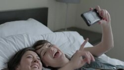 Young women fall back onto hotel bed and laugh at photos on smartphone Stock Footage