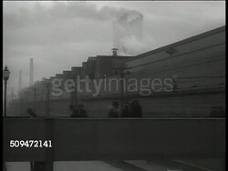 1939: U.S. FORD AUTOMAKER:  EXT WS Parked cars in 'Ford Motor Company' factory lot. VS Workers entering factory. INT VS Workers on assembly flipping car frame, applying car body to chassis. EXT MS Truck of Deluxe Coupes leaving lot. Automobile Instructional Video