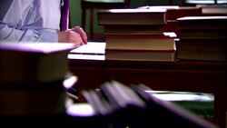 Man wearing spectacles sitting at desk reading books with more books on desk Stock Footage
