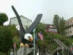 "Bald eagle wooden statue, road tunnel, people, cars, wooden buildings, Alaskan state flag and national flag of USA flying in background, on corner of Front Street, Grant Street and Water Street, Ketchikan, Ketchikan Borough, Alaska." Stock Footage