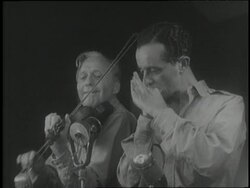 Comedian Jack Benny plays violin with harmonica virtuoso Larry Adler during a USO show. News Clip