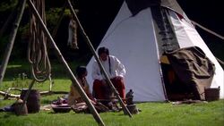 Older male Native American Indian sitting talking to seated child and joined by another male with teepee behind and ropes and dead animal hanging from wooden pole Stock Footage