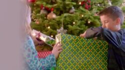 Young boy and girl inspect presents, trade back and forth beneath Christmas tree (dolly-shot) Stock Footage