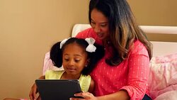 MS Smiling mother and daughter sitting on bed reading book together on digital tablet Stock Footage