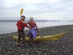 WS Senior couple hugging and looking at view next to kayak on pebble beach / Washington State, USA Stock Footage