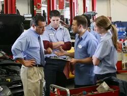 MS Mechanic goes over some details with three other co-workers and manager while standing in front of open hood car / Seattle, Washington, United States  Stock Footage