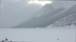 Distant person crosses frozen lake to ice fishing shack, mountains Stock Footage
