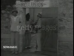 1940: FRENCH GUIANA (Iles du Salut): Doorway sign: 'Reclusion', Guard standing in doorway, pushing prisoner back. LS INT Long line of doors w/ trustee filling extended cups from doors w/ ladle of food. CU Male face looking out of door square. Instructional Video