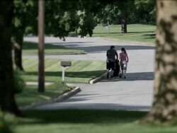 Young couple walking and pushing baby stroller on suburban street.  Framed through out-of-focus flowers and trees in the foreground. Stock Footage